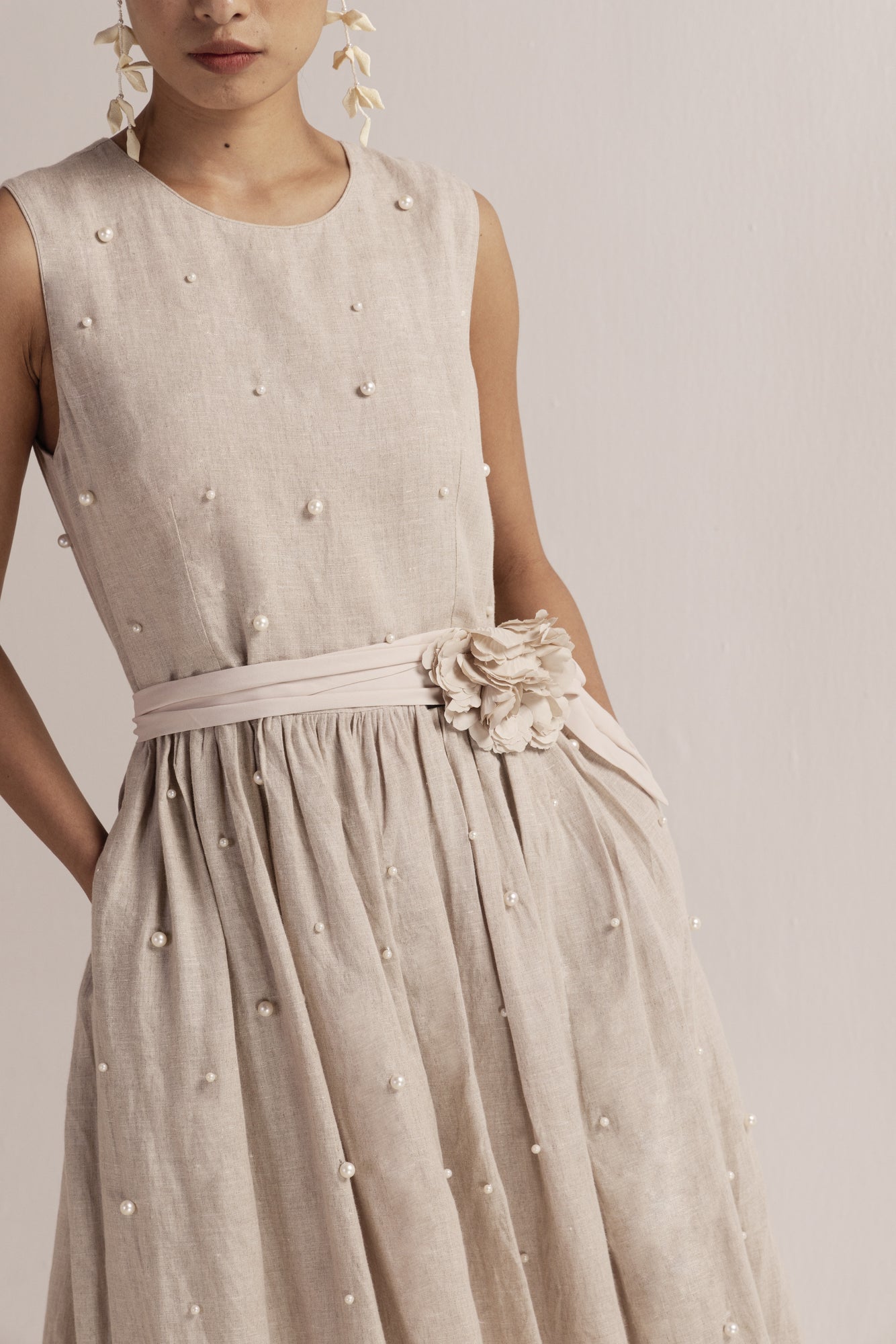 Beige linen mid-length dress with pearl embellishments, sleeveless design, round neckline, and soft waist gathers for a refined silhouette